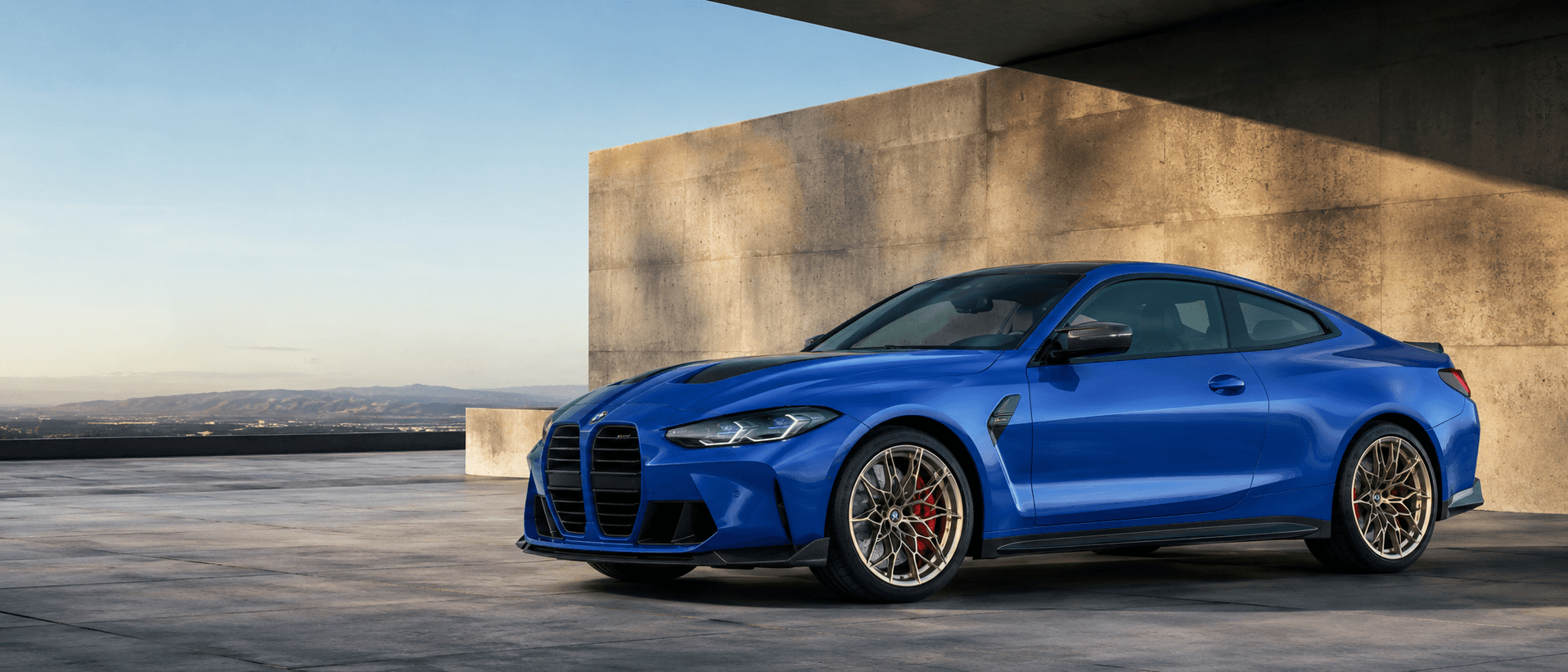 Beton mimari önünde mavi BMW M4 CS yan ve ön üç çeyrek görünümü
